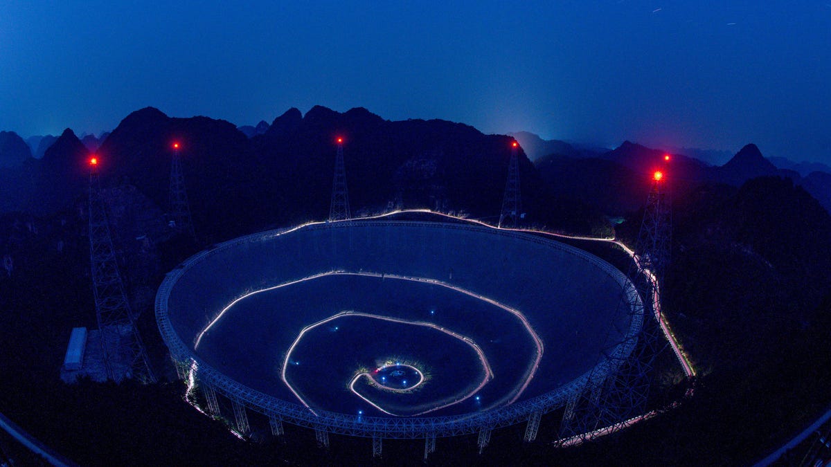 A large antenna dish, cut into a mountainside, is illuminated by lights spiraling toward the dish's center