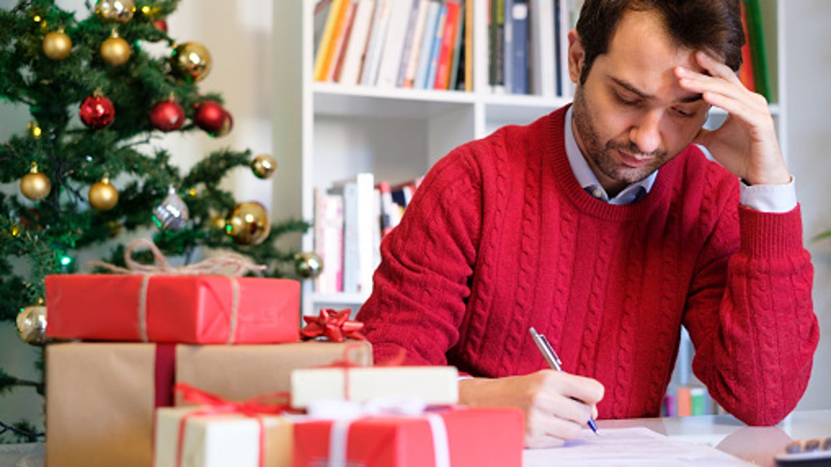 Man stressed while writing next to a stack of gifts with a Christmas tree in the background.