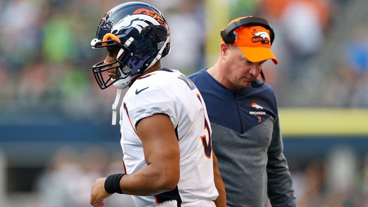 Quarterback Russell Wilson looking off to the left in white Broncos jersey. Head coach Nathaniel Hackett is in the background looking dejected.