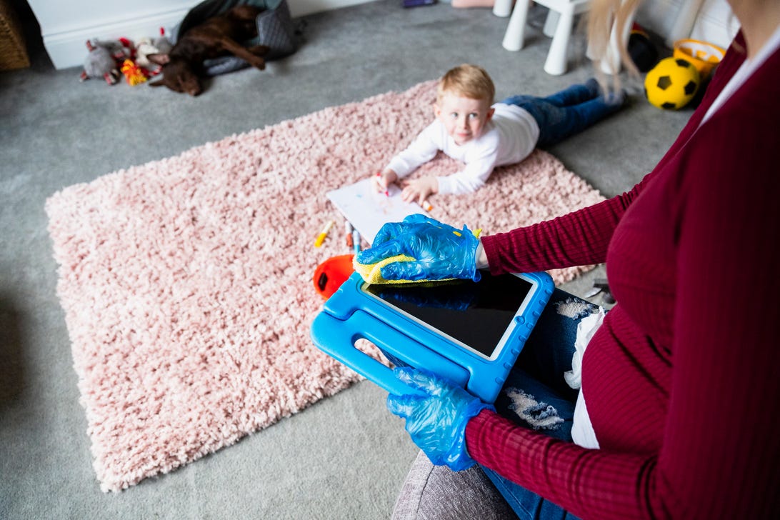 Person in rubber gloves cleaning a toy's screen with a baby in the background