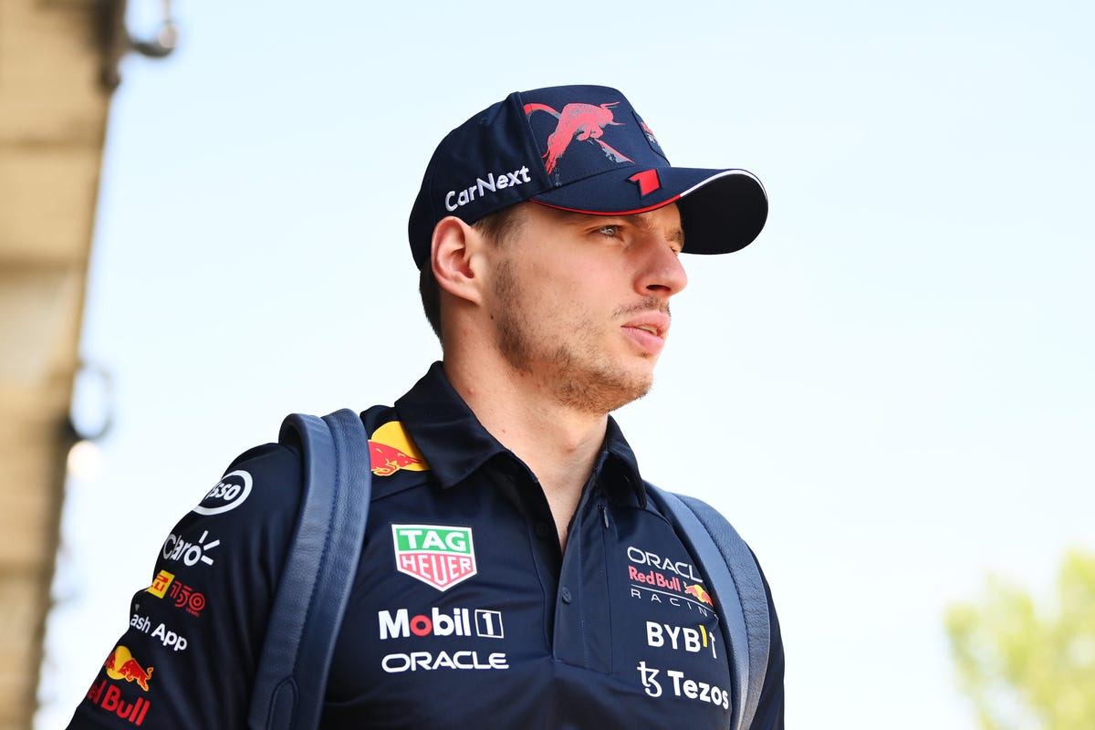 Max Verstappen, dressed in a Red Bull uniform, hat and backpack, stares into the distance.