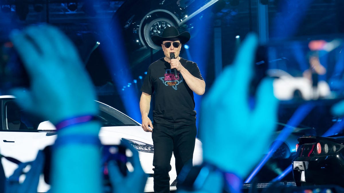 Elon Musk in black T-shirt, sunglasses and cowboy hat, speaking into a handheld mic