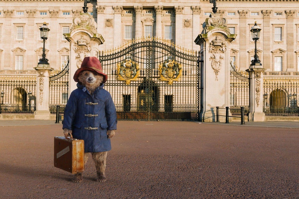 Paddington Bear, carrying a suitcase, stands outside Buckingham Palace