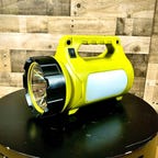 The LE Rechargeable LED Camping Lantern sits atop a black table in front of a wooden wall. It's a large-sized flashlight that doubles as a convenient lantern for camping trips.