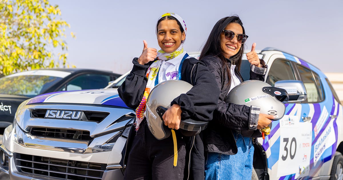 Rally Jameel drivers in front of an Isuzu race car, holding their helmets and giving a thumbs-up