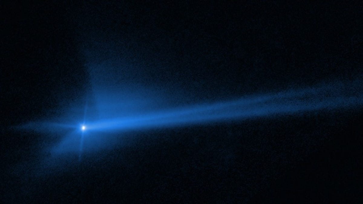 Hubble image of asteroid Dimorphos after DART spacecraft impact. The asteroid look like a bright dot with hazy blue debris emanating from it and stretching out behind into a split tail.
