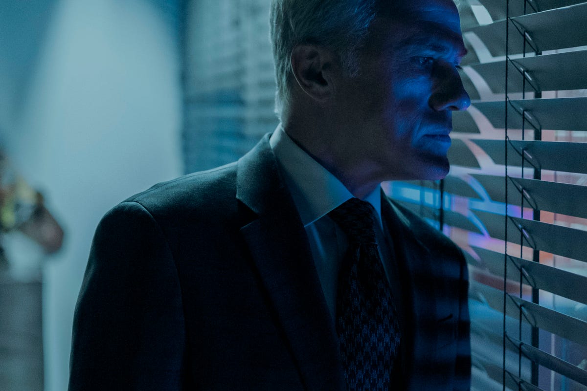 Christoph Waltz in a suit peering through the blinds of an office in shadow