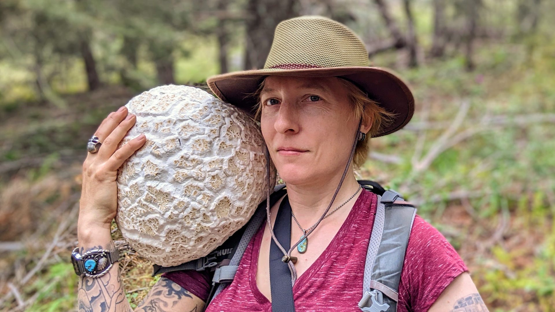 A mushroom forager in a red shirt and brown hat holds a massive crackled-looking round puffball mushroom on her shoulder.
