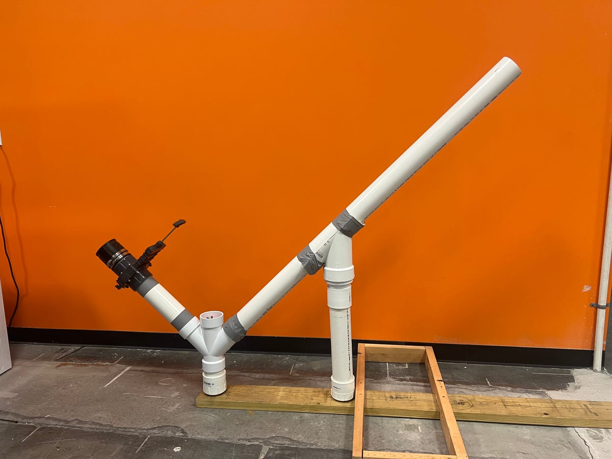 A custom-built air cannon made from PVC pipe sits atop a wooden frame. It's shaped like a big checkmark. We stick a leaf blower's nozzle into the bottom end and rev it up, then pull a handle to open a valve, which lets all of that air power propel a weight out of the top.