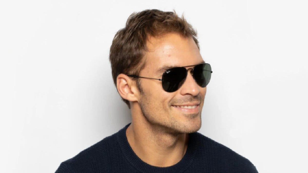 A male wears a pair of Ray-Ban RB3025 Aviator sunglasses while smiling against an off-white background.