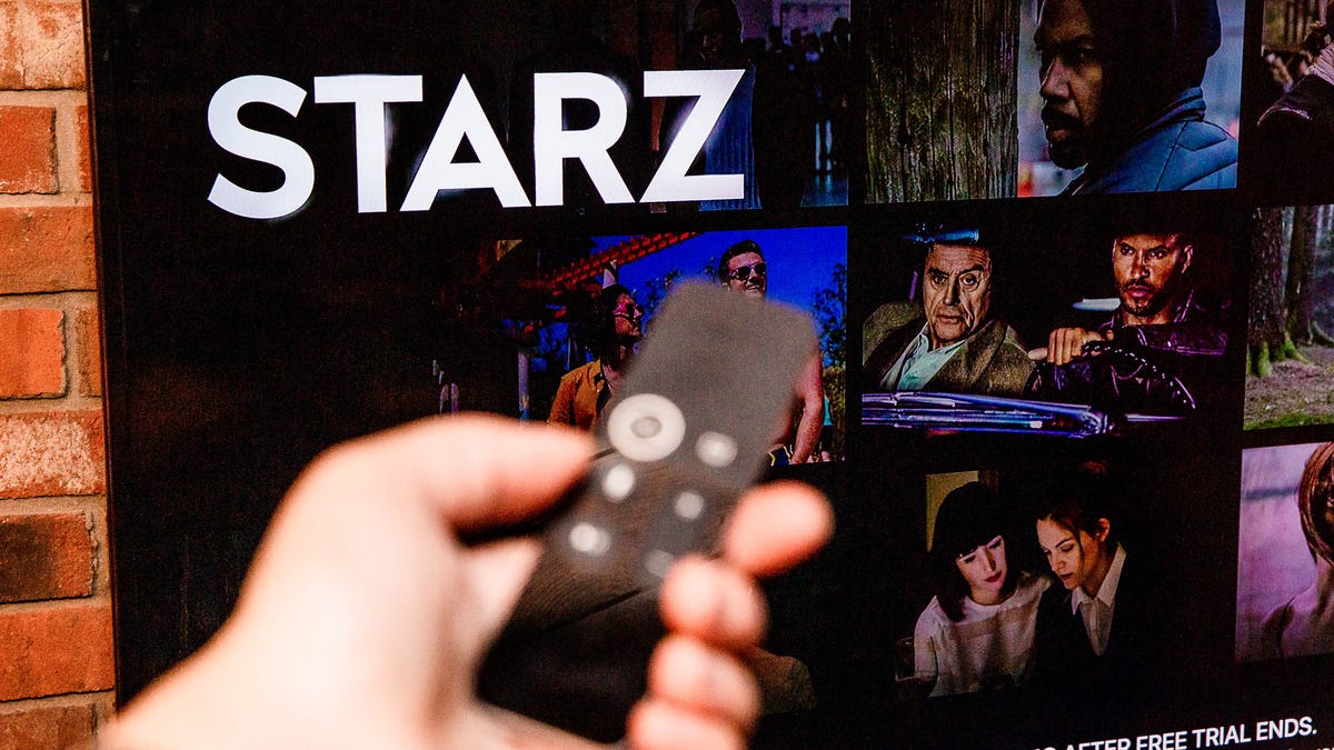 A hand holding a TV remote in front of the Starz streaming app on a flatscreen TV.