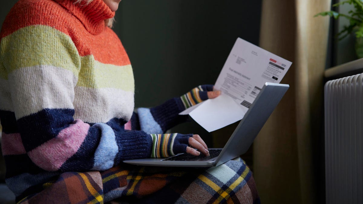 A person in an orange, green, white and blue sweater holds a power bill in one hand and works on a laptop with another hand.