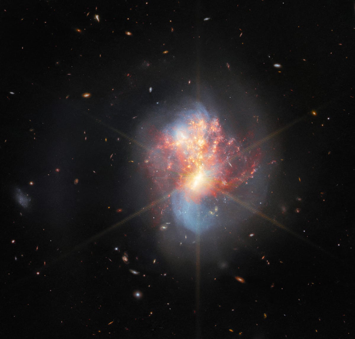Against the background of space, lots of tiny spots represent giant galaxies across the universe. At the center is a scene of two merging galaxies, which looks like a swirls of pink, red and blue blurs. The JWST's iconic diffraction spikes are seen, too.