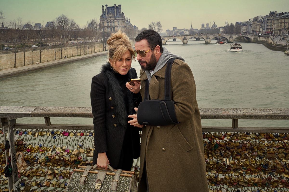 Jennifer Aniston and Adam Sandler looking at a phone on a lock bridge in France