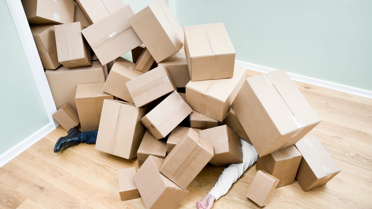 a person lies on a wooden floor covered by a tower of cardboard moving boxes that has collapsed