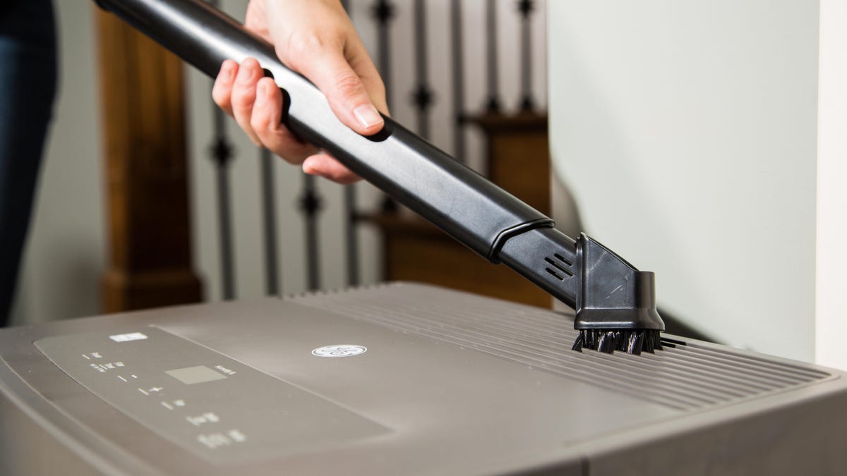 A person uses a wand attachment on a vacuum to clean a dehumidifier