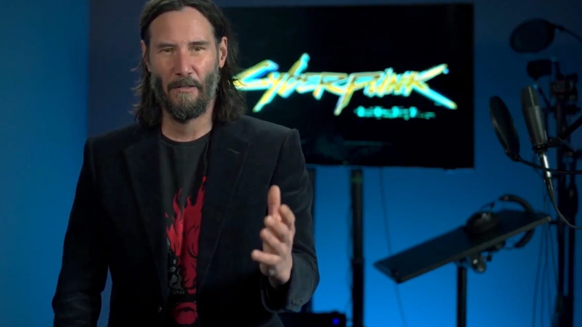 keanu reeves talking to the camera with a cyberpink 2077 logo on a display behind him