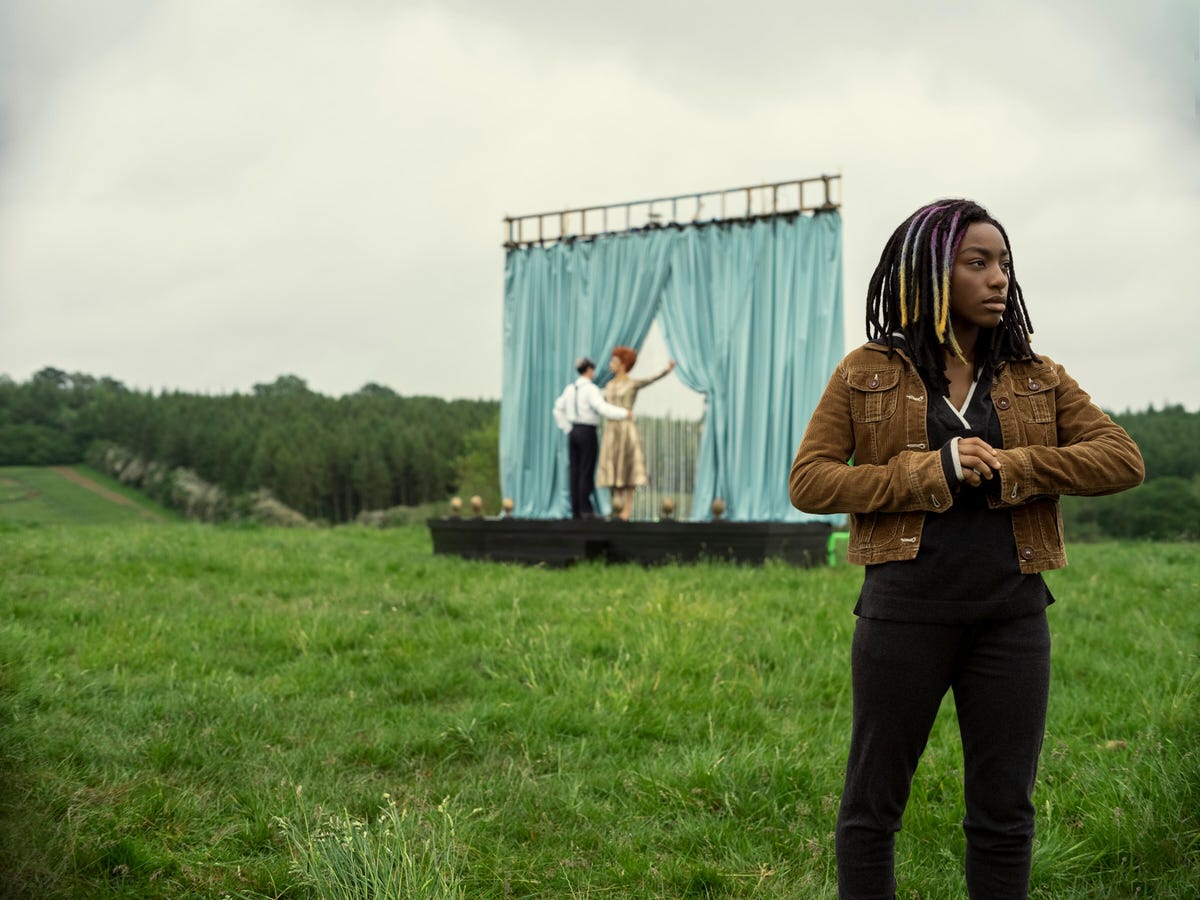A young Black woman stands in a field where two people dance in the distance on a strange theatre stage.