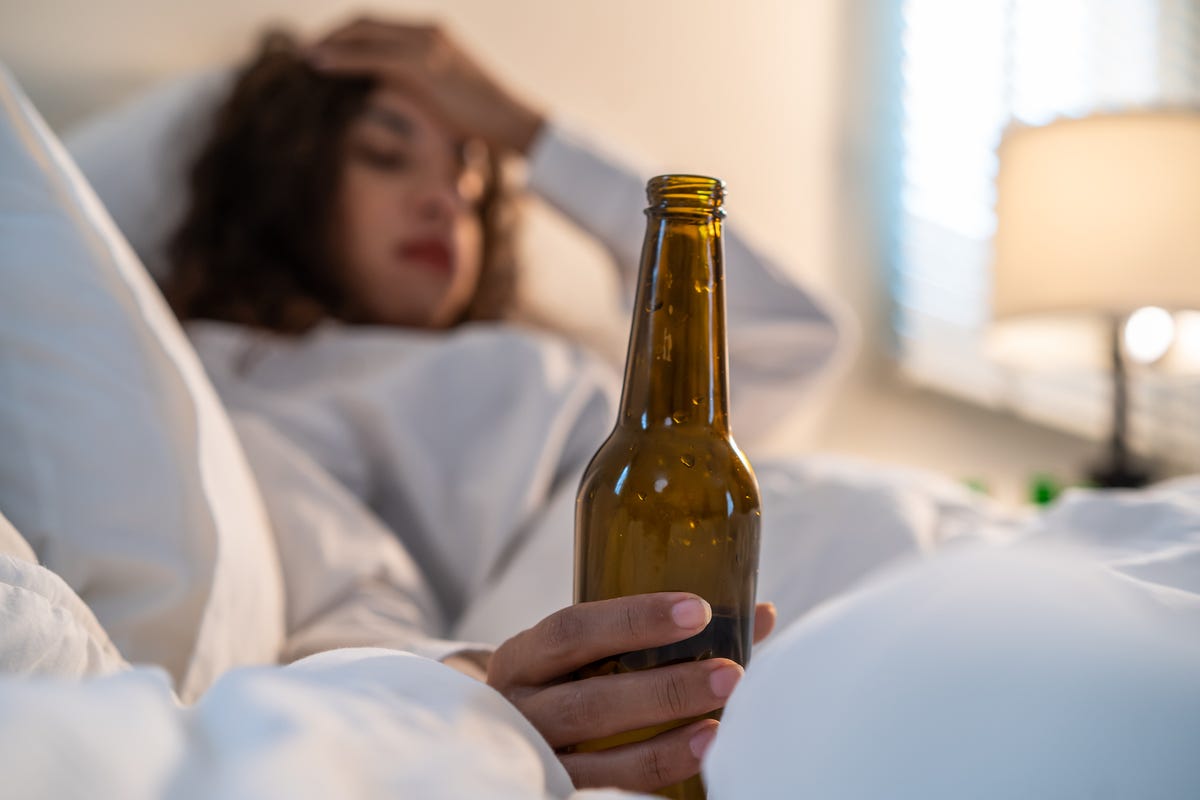 Latino woman holding beer bottle in one hand and head with the other while drinking in bed