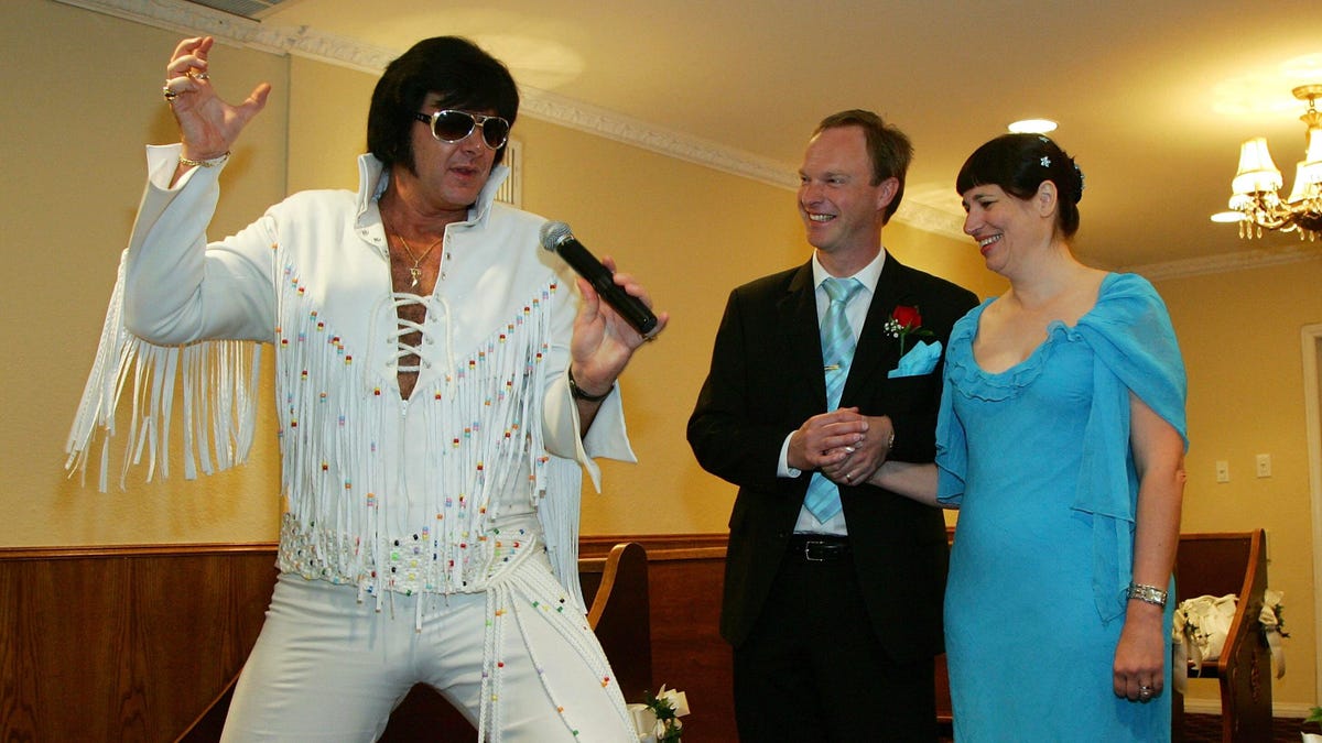 An Elvis impressionist in a white suit with fringes poses with a man and woman dressed for their Las Vegas wedding