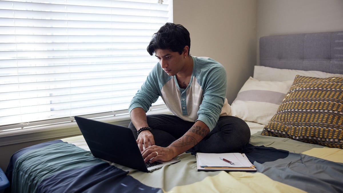 A young person using their laptop on a bed with a notebook by a shaded window.