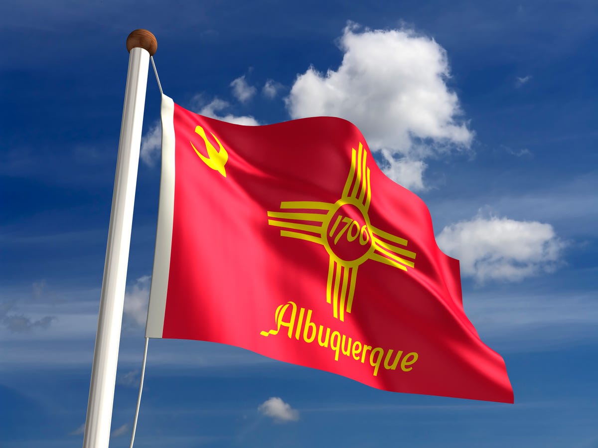 Red and yellow Albuquerque city flag on a flag pole with blue sky and puffy clouds behind it.