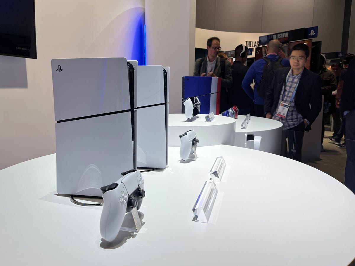 Two white PlayStation 5's with a blue, silver and red PlayStation 5 int he background
