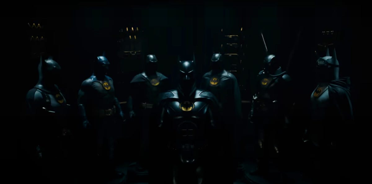 A collection of seven Batman costumes are displayed in a dark room in the Flash.