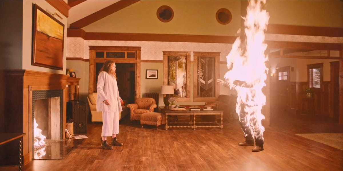 A still from Hereditary. A woman stands before a man who is on fire.