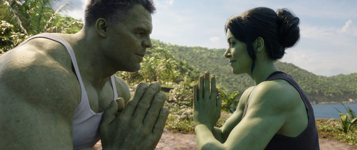 She-Hulk, a woman with green skin and superpowers, adopts a yoga pose and smiles at the bigger but equally green Incredible Hulk.