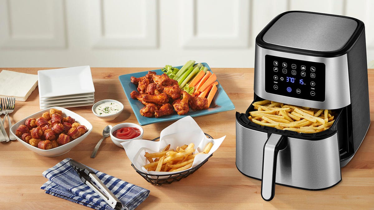 A stainless steel Insignia air fryer rests on a wooden surface with wings, fries and other foods in and around it.