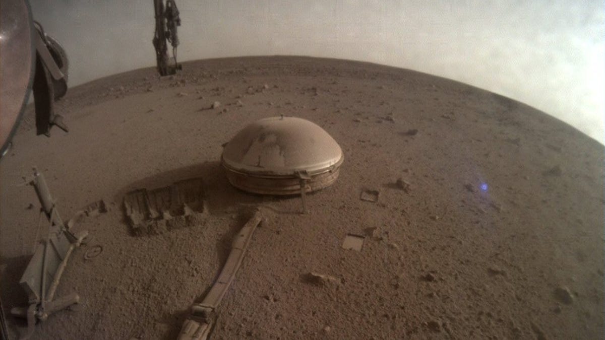 Dusty Mars landscape with curved horizon, parts of the InSight lander visible and a dome-covered seismometer.