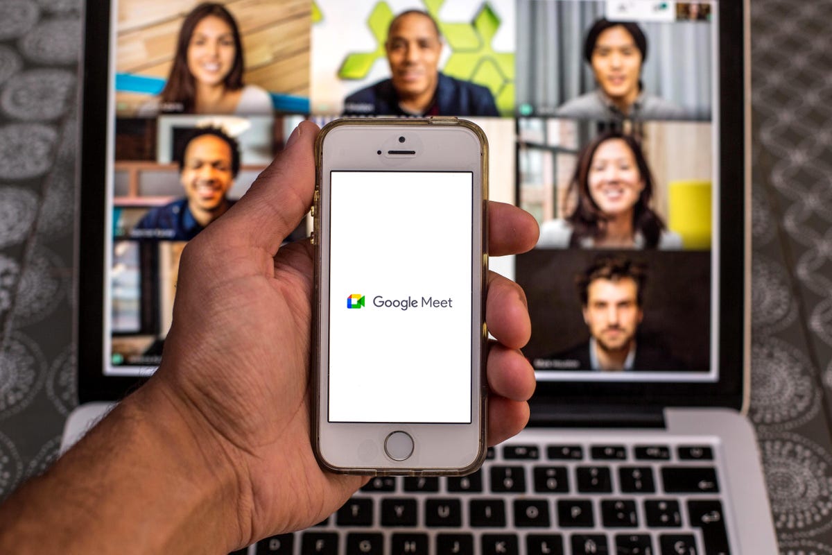 iPhone showing the Google Meet logo in front of a laptop showing the faces of others in a video conference