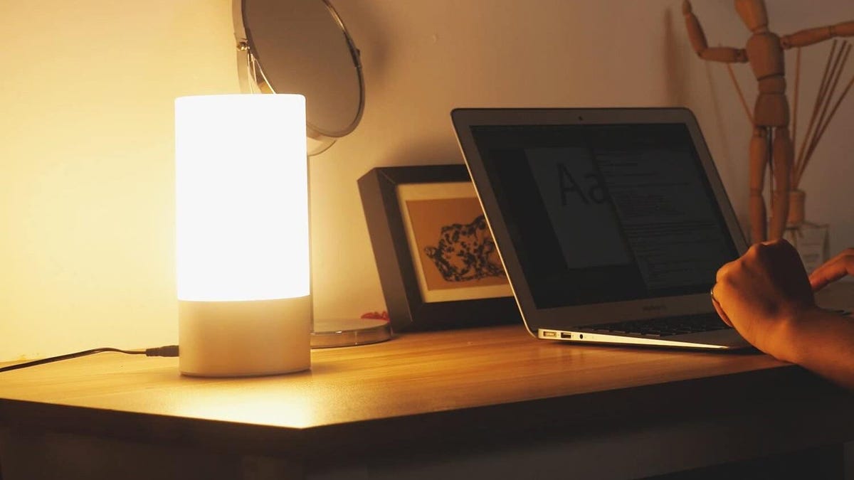 A lit Hifree table lamp on a desk next to someone working on a laptop.