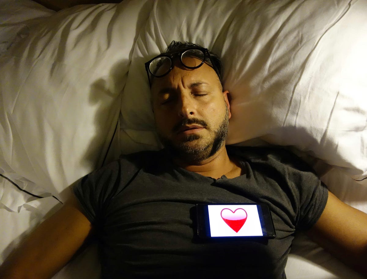 Un homme allongé dans son lit avec son téléphone sur sa poitrine.  Le téléphone affiche un emoji cœur