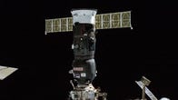 Russian Progress 82 cargo craft, a dark cylinder with outstretched solar panels like a cross, is connected to a dock on the ISS.