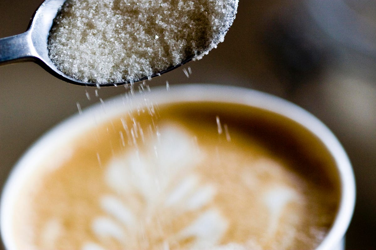 A spoon of sugar being poured into a latte. Latte has a pattern with milk and espresso.