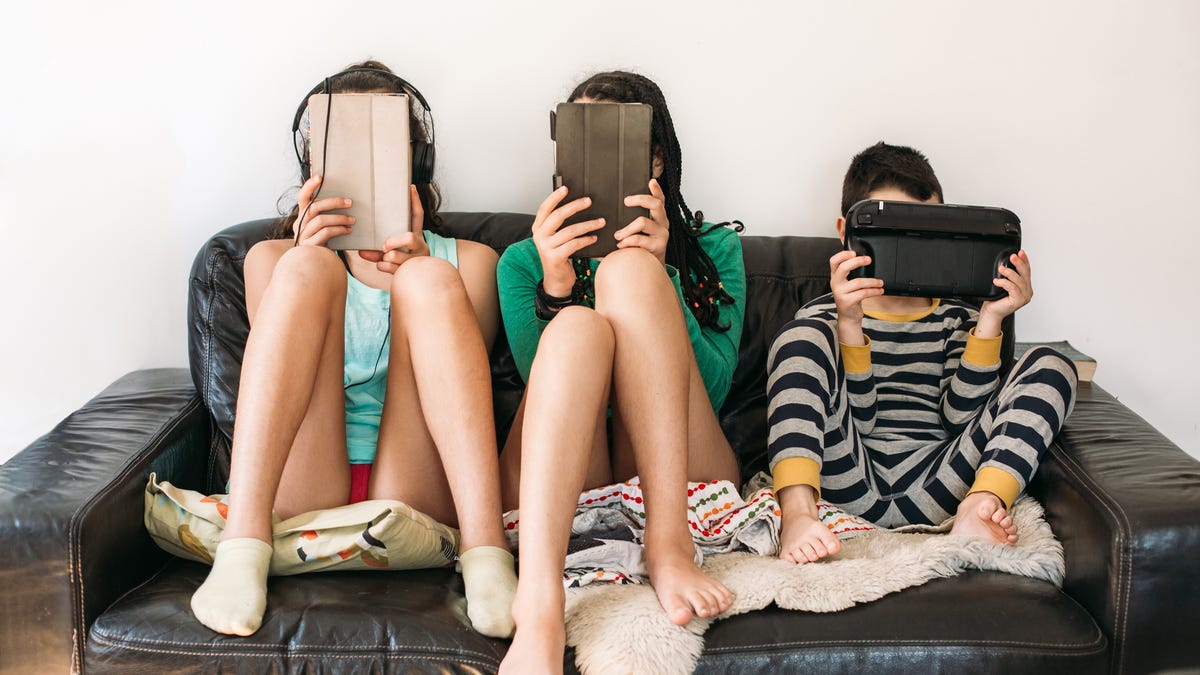 Three children sitting on a couch holding tablets up to their faces