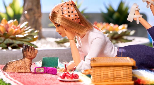 Barbie lying on picnic blanket with a dog, a book, a flip phone and some cherries