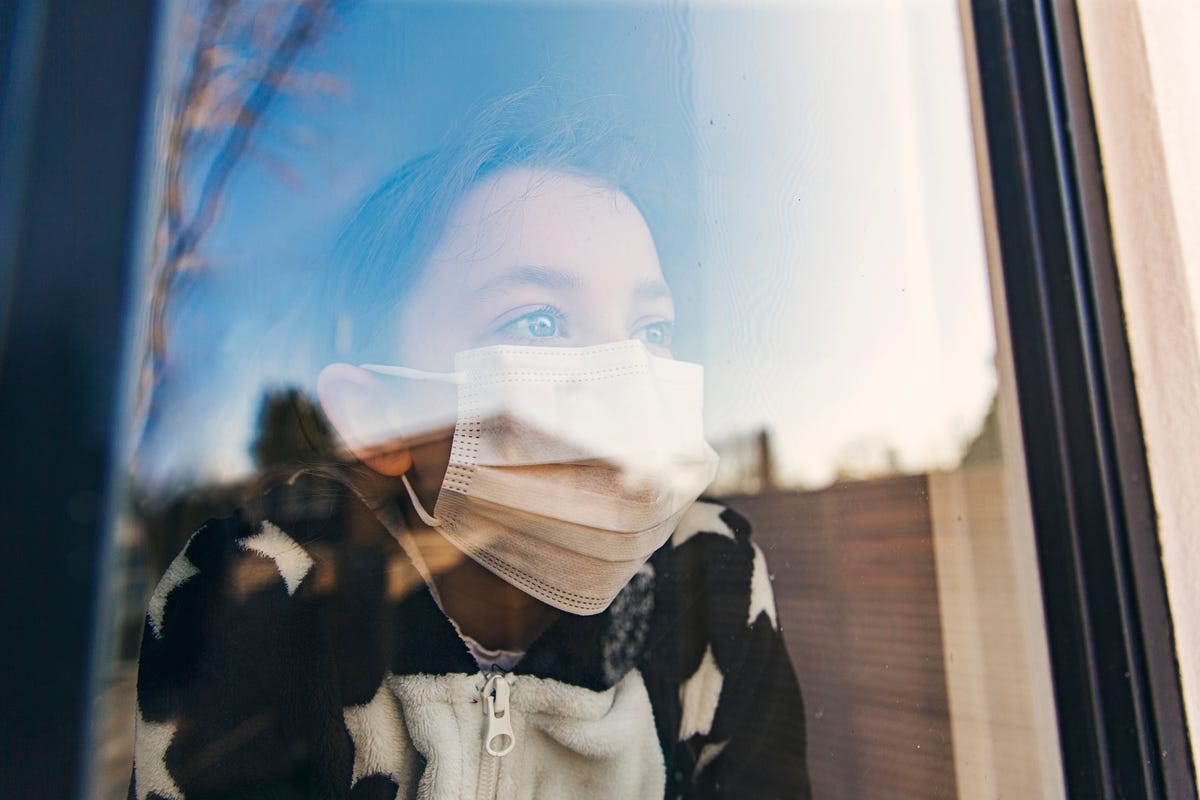 A child looks out a window wearing a mask