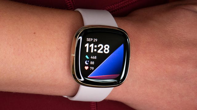 A wrist wears a Fitbit Sense with a brightly-colored, digital clockface displaying the time and a pink band.