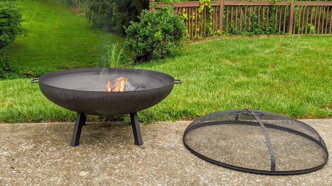 The large Hampton Bay fire pit on an outdoor patio with a small flame inside and the cover on the ground next to the fire pit.