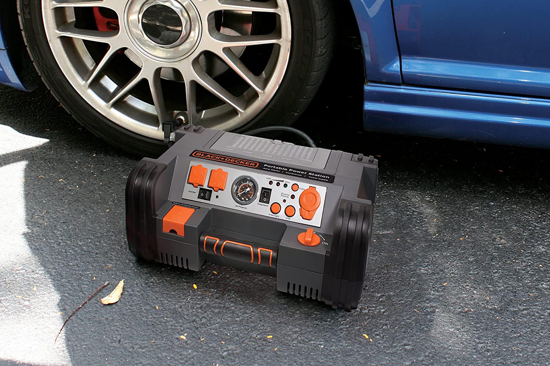 An orange, black and grey Black & Decker jump starter on the ground next to a car.