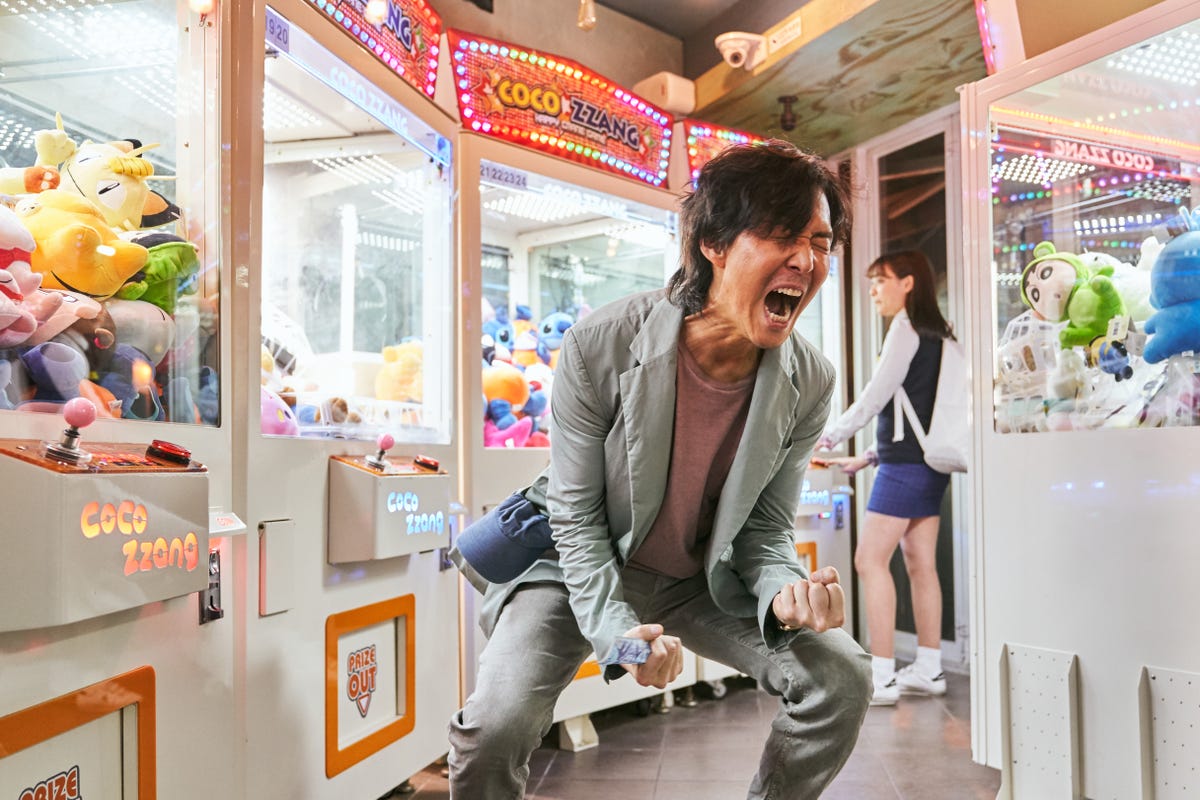 Character Seong Gi-hun screams with clenched fists inside a colorful, brightly lit arcade.