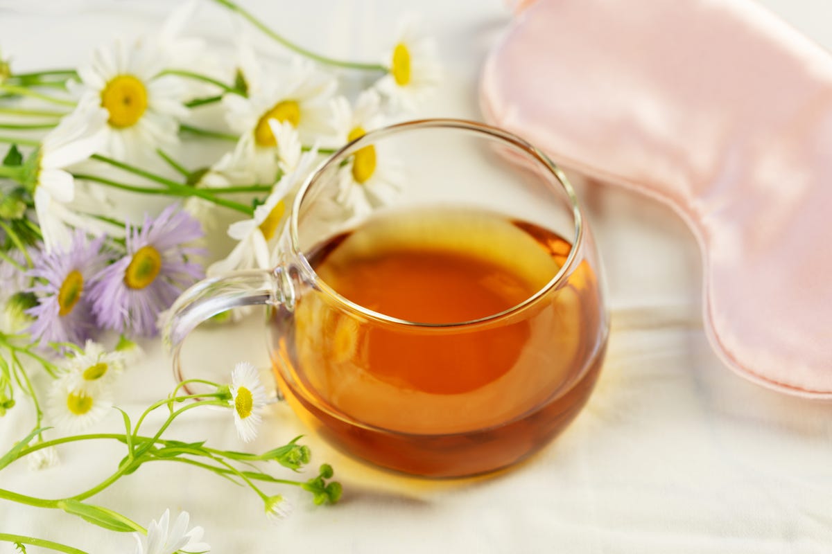 Glass cup of tea, sleep mask and flowers on a white bed sheet.