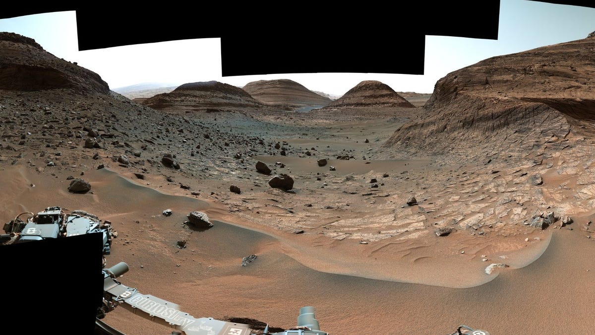 Parts of the Curiosity rover visible near the bottom of a Mars landscape image shows rounded hills, sand and rocks.