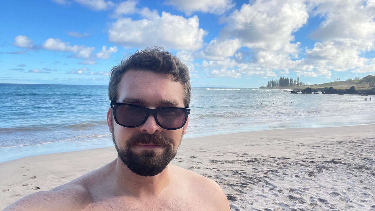 A man in sunglasses takes a selfie with a Maui beach behind him.