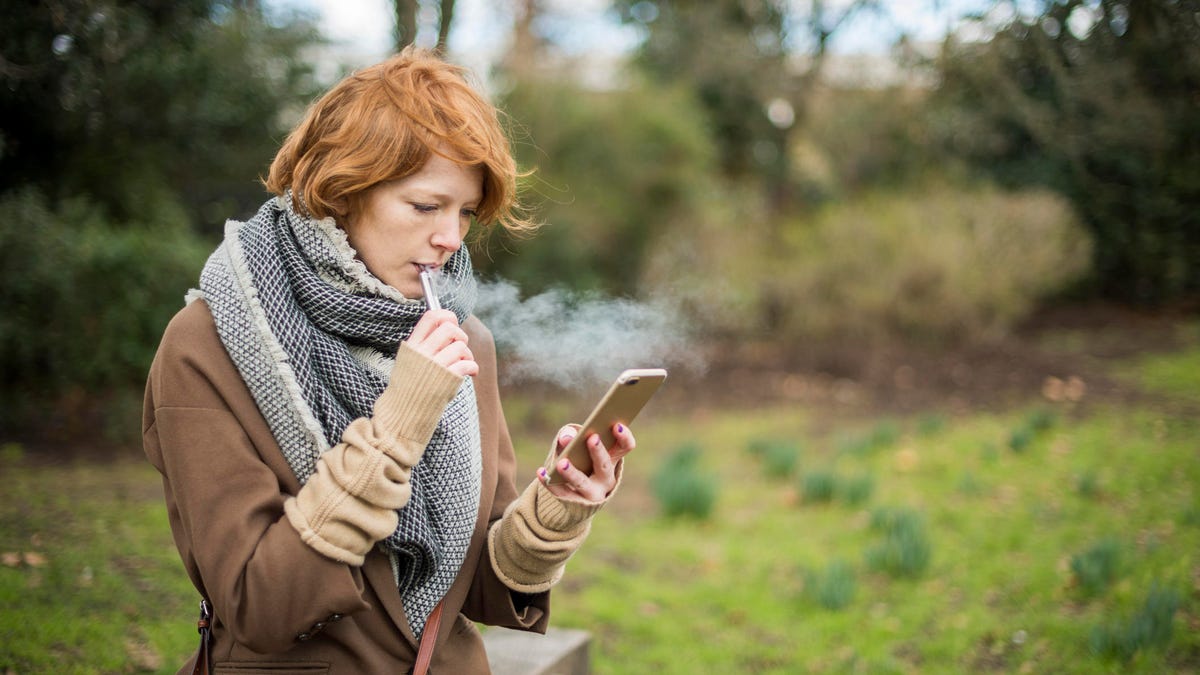 Red haired woman using an electronic cigarette and a smart phone in the countryside