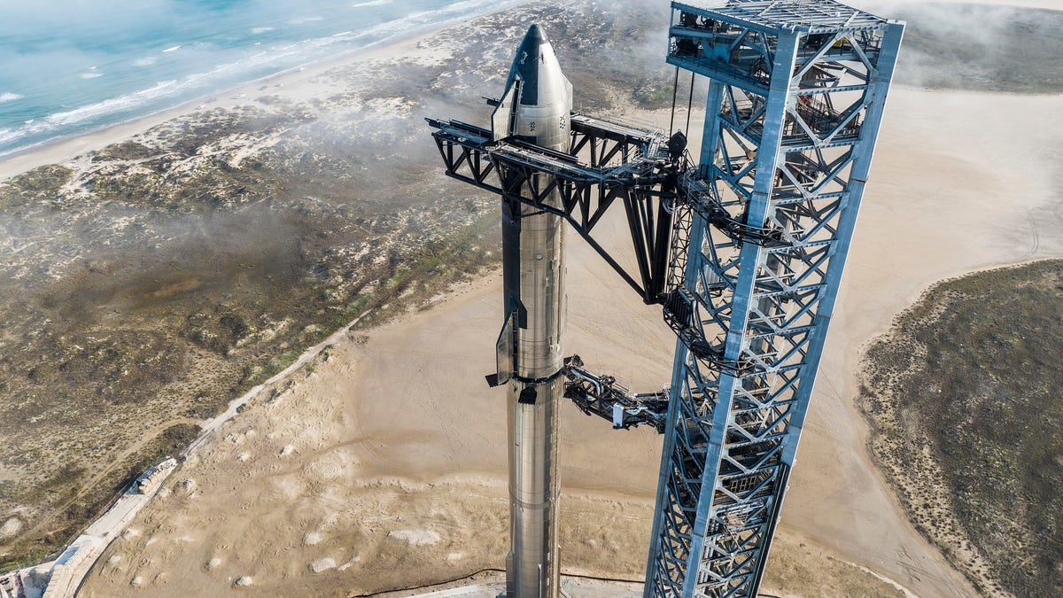 Shiny side of SpaceX's Starship spacecraft angled toward the camera, stacked on a long shiny rocket with launch tower beside it and Texas in the background.