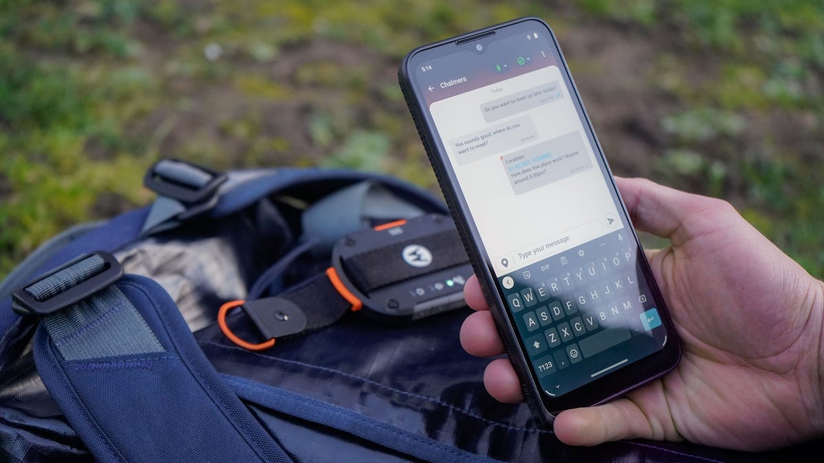 In a wilderness setting, a smartphone with a messaging app open is positioned above a backpack and the Satellite Link device.
